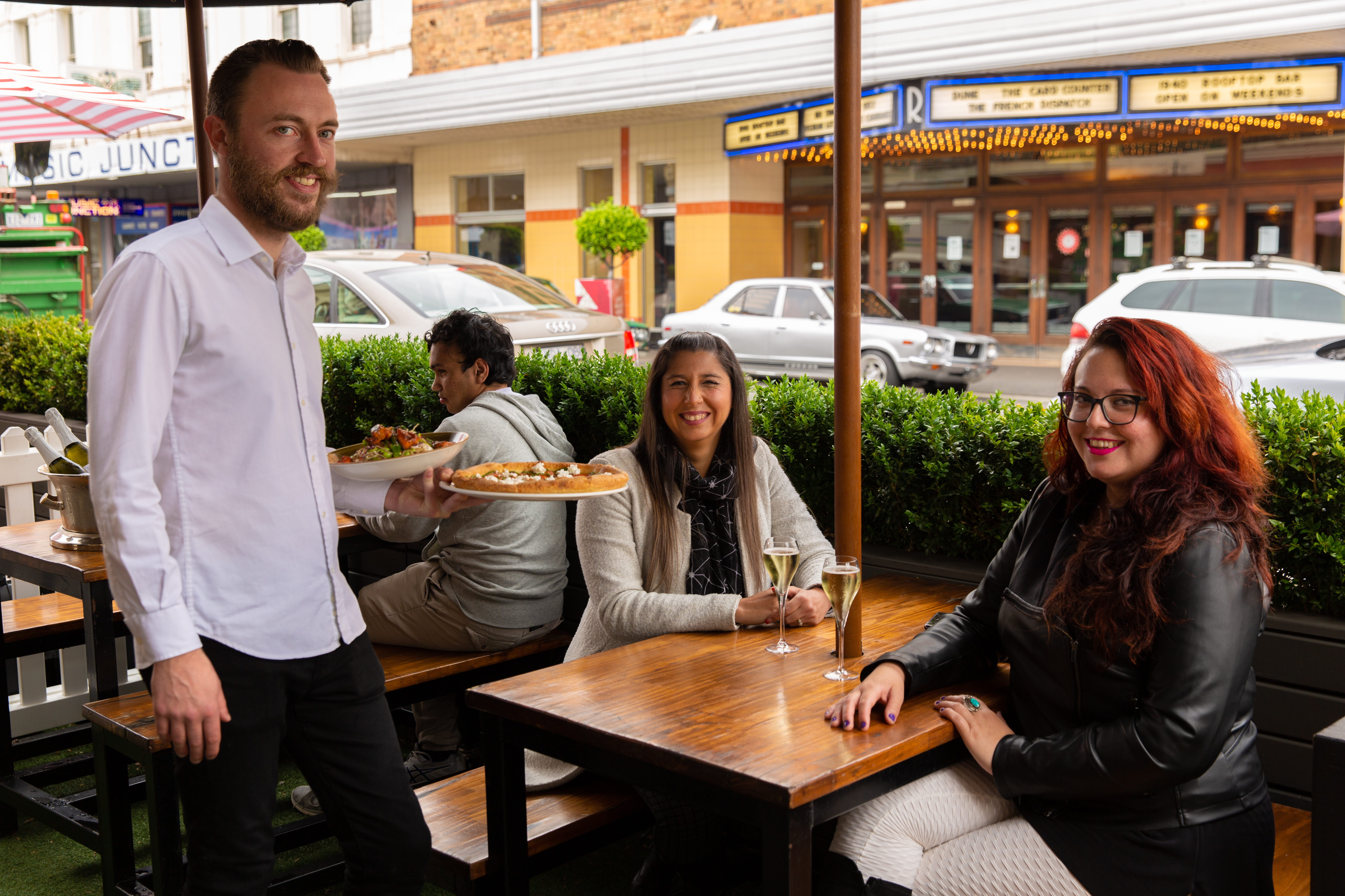 Three people around a table that is on the side of the road in a parklet area bordered by plants between them and the road