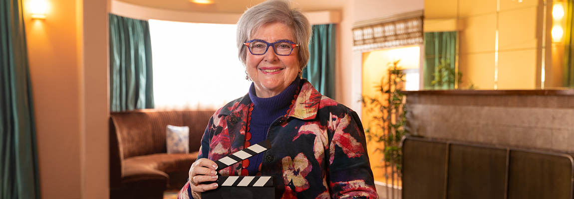 An older person standing inside smiling and holding a film clapper board