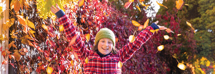 A young person wearing warm clothes on a sunny street surrounded by colourful autumn leaves