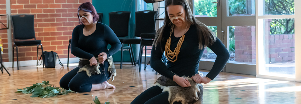 Two Indigenous women with face paint and possum skirts kneel on a wooden floor