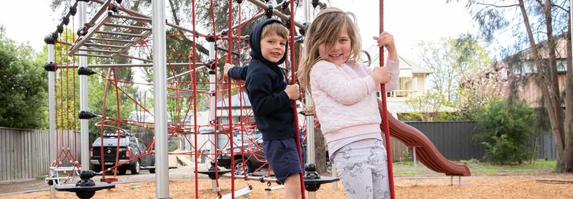 Two preschool children hang off an outdoor rope climbing structure