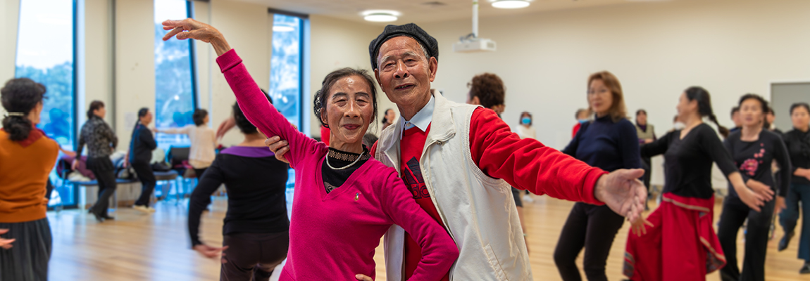 An older Asian couple dance in a room full of other people dancing