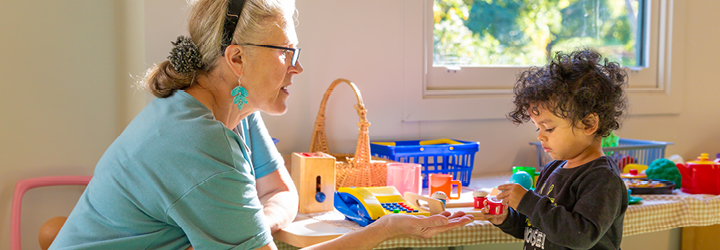 An older woman with a ponytail plays at a Lego table with a preschooler