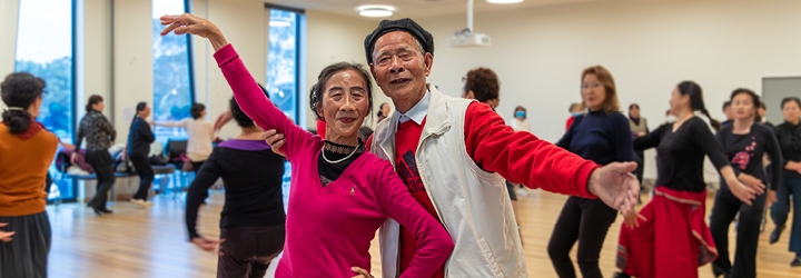 An older Asian man and woman hold their arms out in a dance movement at a dancing class