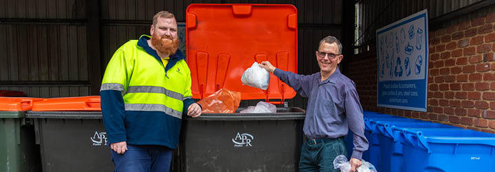 a man in high vis jacket and a man in a collared shirt dispose of soft plastics in a skip