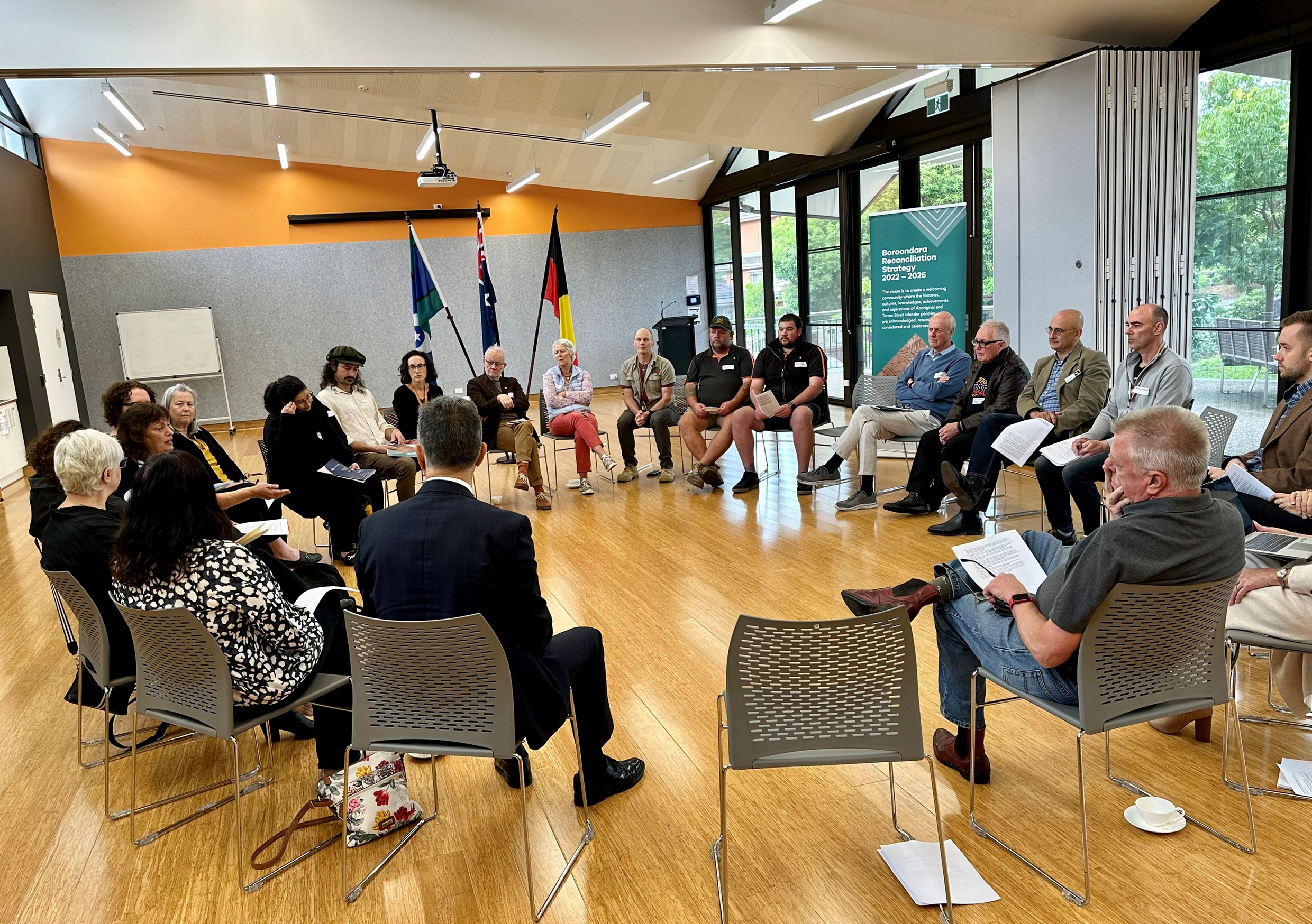 A group of people sitting indoors in a circle on chairs, with the Australian flag, Torres Strait Islander flag, and Aboriginal flag behinf them.