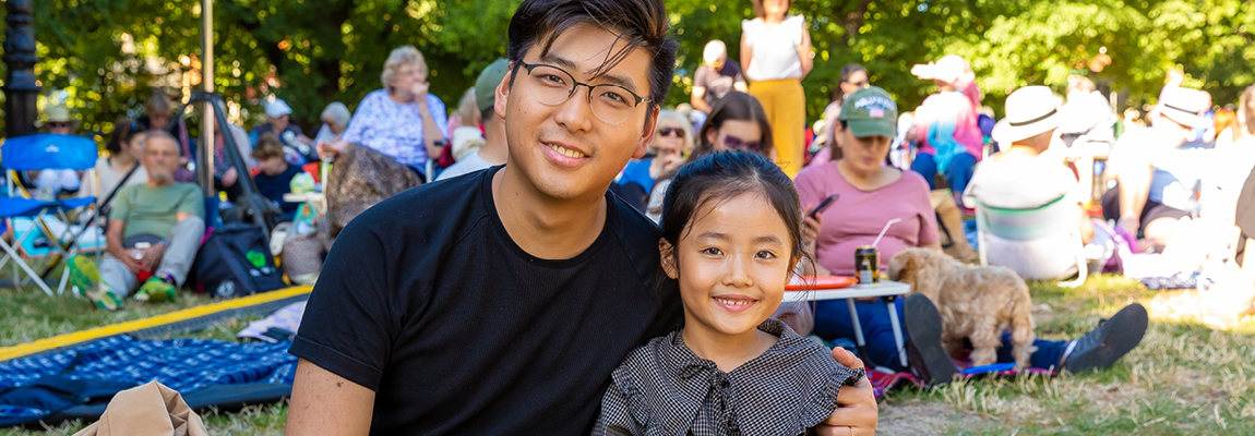 Man and a young girl looking at camera with people picknicking in background in a park