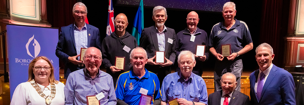 A group of 11 people standing together at an awards ceremony, some with plaques