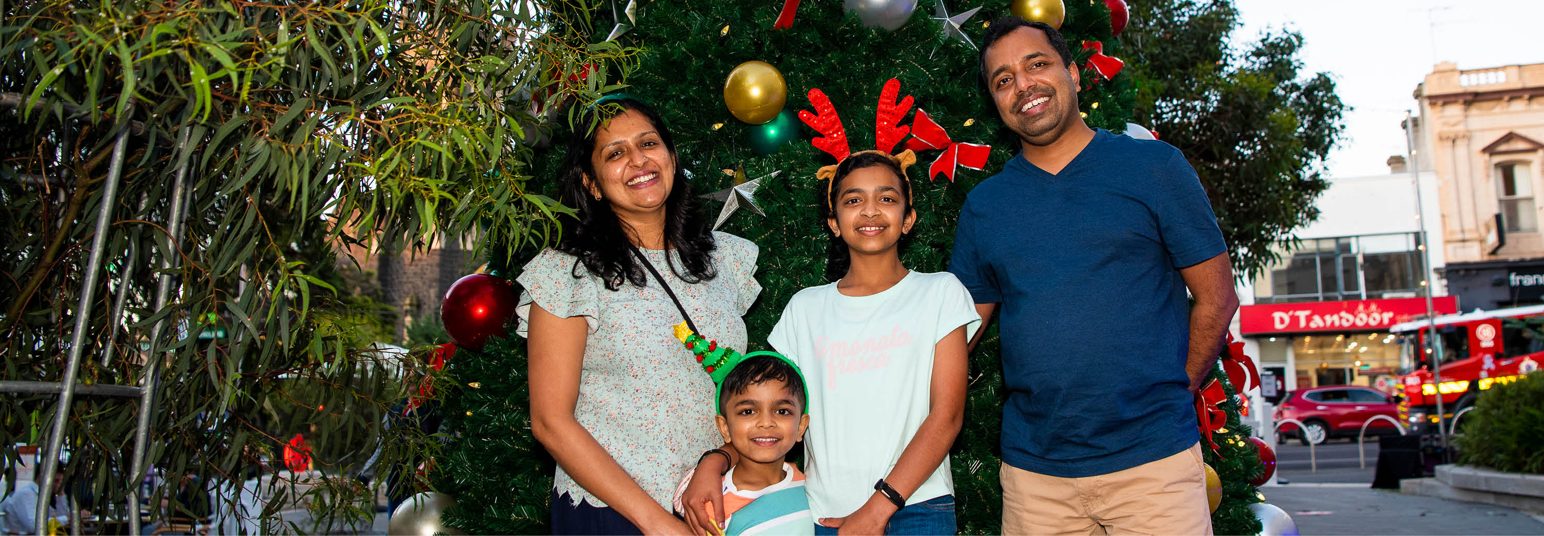 An Indian family smile in front of a Christmas tree with a Tandoor restaurant behind it