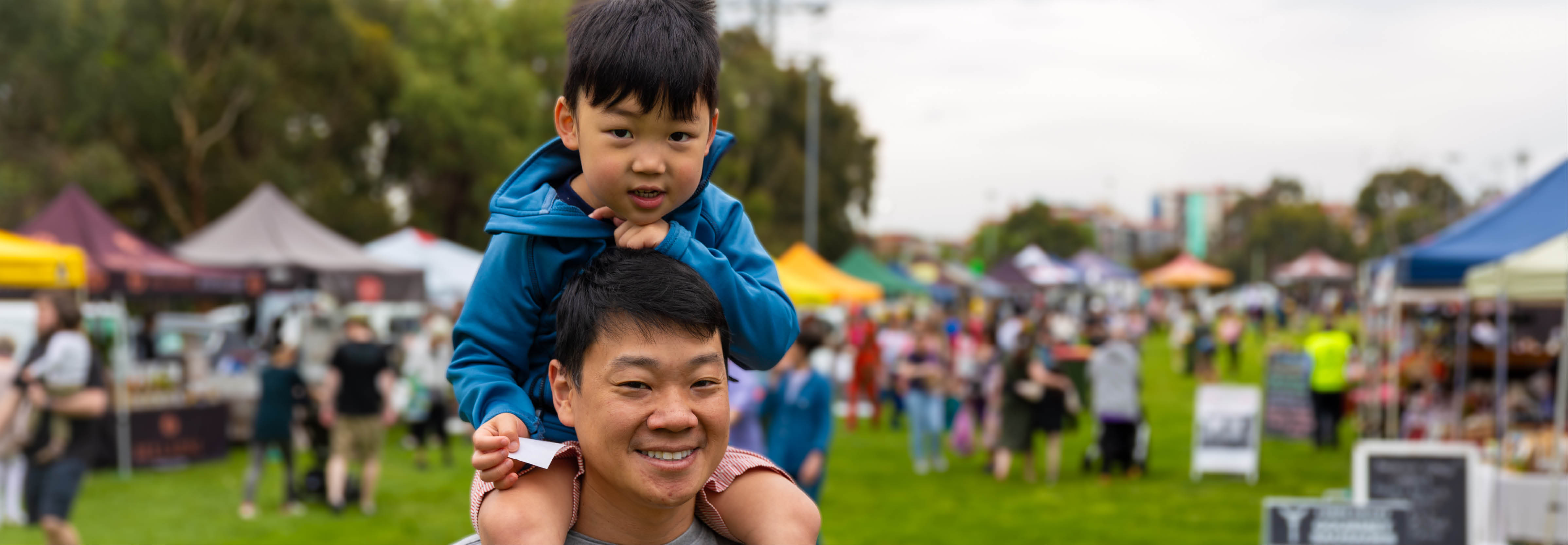 a man carries a young boy on his shoulders at an outdoor market with stalls and tents