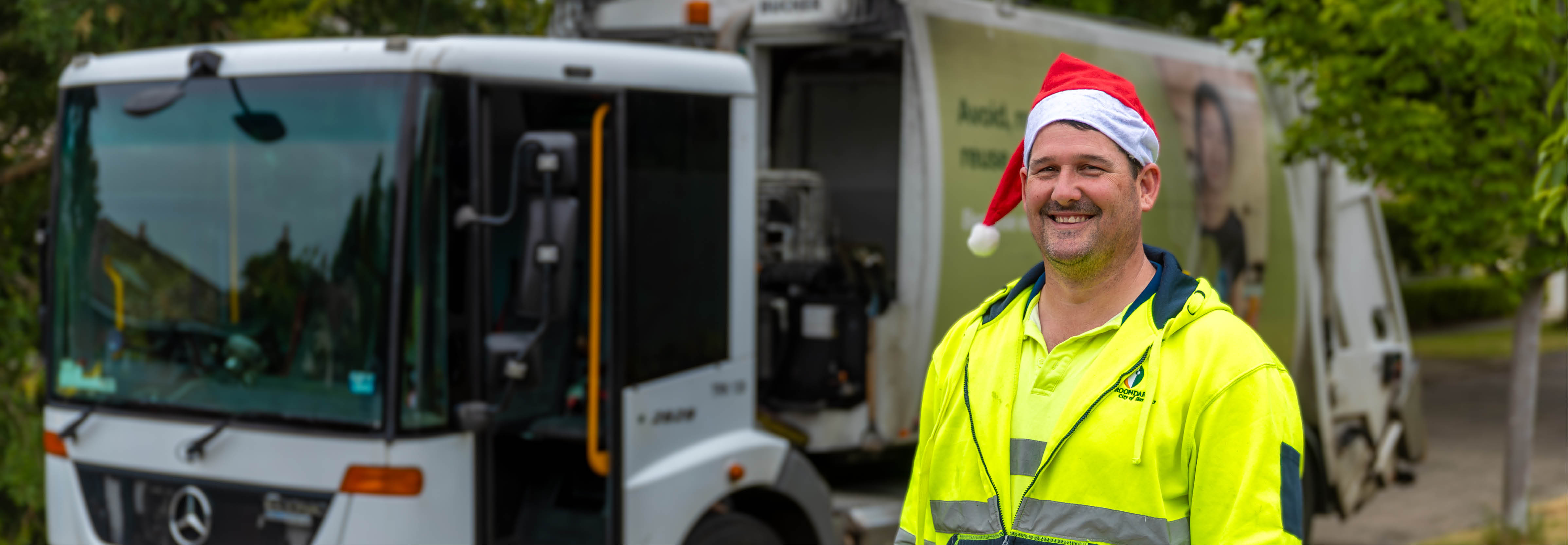 a man in high-vis shirt and santa hat smiles beside a garbage truck