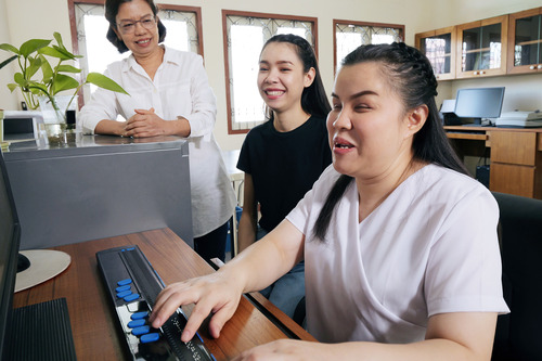 3 people working at a computer. 1 person is using a refreshable braille display