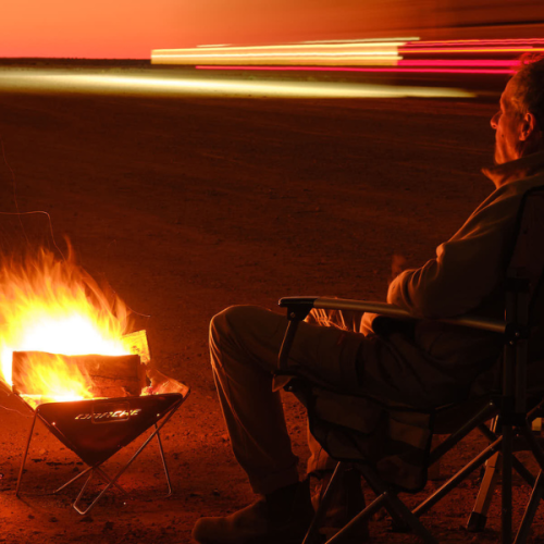 A man sits in a camp chair beside a campfire in a small drum