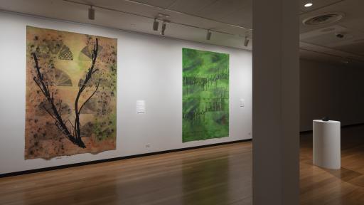 The exhibition space for Above the Canopy, showing a large gallery room with two pieces of art on the wall, one showing a large branch with fire danger rating signs behind it, and the other a mottled green canvas with a graph of rising temperatures shown