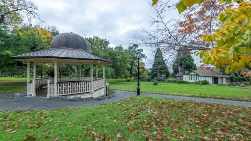 Rotunda with cream-white balustrades and grey dome-shaped roof, surrounded by a footpath and grass area. There is a lamp post in the centre and fallen leaves on the ground.
