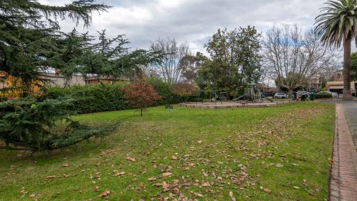 Grass area bordered by hedge to the left and walking path to the right. There are fallen leaves are on the ground and a playground and parked cars in the distance.