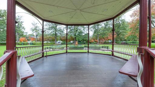 Inside view of round outdoor rotunda located in grass area, with trees in the distance. The columns and bench seats are dark red and the roof and balustrades are white.