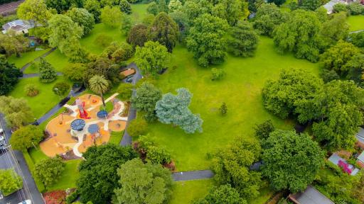 Aerial view of grass area with many scattered green trees. There is a large playground with surrounding footpaths to the left.