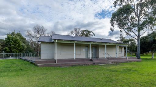 Single-storey cream white weatherboard building with sloped corrugated roof, on the edge of a sportsground. There is a tall tree to the right and small trees in the background.