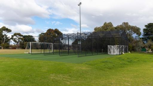 Cricket nets with 5 lanes divided by tall black fences. There are tall trees and a light post in the background.