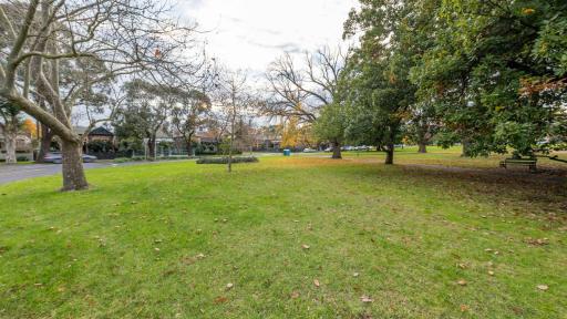Large grass area with fallen leaves and scattered mature trees. There is a park bench and walking path to the right and a line of houses in the distance.