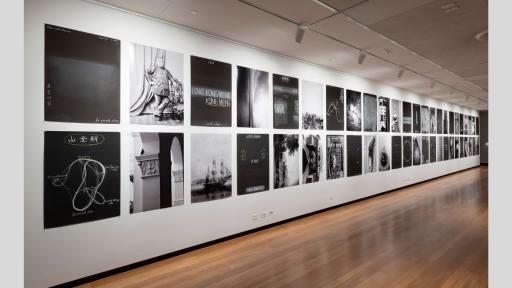 An installation in a white-walled art gallery of 49 black and white photographic prints and chalk drawings, each measuring 100cm by 70cm. The photos variously depict architectural elements, ships, street scenes, historical figures, neon signage, and chalk scrawls.