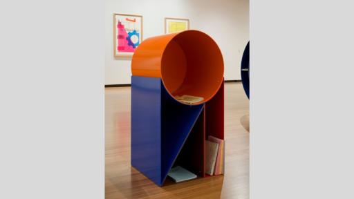 A metal sculpture, about a metre tall, standing in a white-walled gallery. It consists of a hollow orange cylinder, on top of a hollow blue and orange straight-edged structure. A collection of small books and magazines are placed in the hollows.