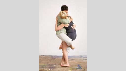 A photograph of a woman and a man in a tender, standing embrace, in front of a white wall. Their right legs are wrapped around each other’s thighs, and their arms are wrapped around each other’s torsos. The woman has her head turned away from us, and the man’s eyes are closed.