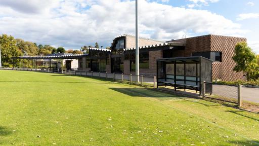 A modern bruck building and undercover area next to a sports oval. The sports oval has wire fencing. 2 players seating/stalls are on the oval boundary.