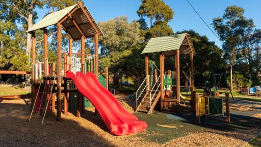 Playground with two small climbing features with slides, surrounded by a grass area and tall trees. There are swings in the background and tall trees in the distance.
