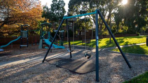A playground featuring a swing seat, 2 slides and climbing features. There is a grass area and a wooden seat in the background.