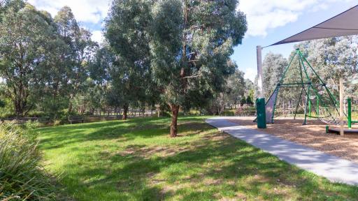 Small grass area with two medium-sized trees, sloping down to two park benches, a wooden fence and foot bridge to the far left. There is a footpath and part of a playground to the right.