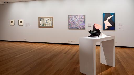 A plinth with an abstract geometric black and pink sculpture on it. in the background 4 artworks are hanging on the wall. The closest artwork is an abstract painting in black and pink, like the sculpture.