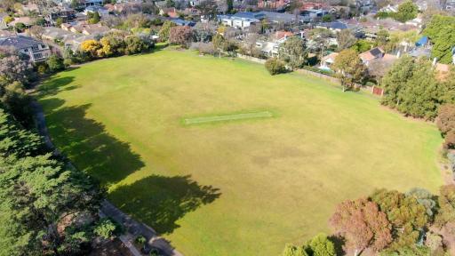 A overhead view of a large sportsfield surrounded by large trees. A green cricket pitch is in the centre.