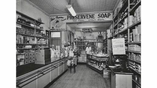 Black and white photograph of a grocer shop. Shelves lined with tinned and bottled goods line the walls of the store and there is a long table with a cash register. Three staff are in the background, one behind a counter at the back of the shop and two near the register partially concealed by product displays.