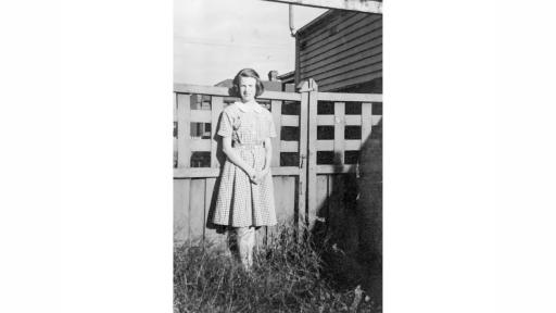 Black and white photograph of a woman with chin length hair style into big waves and wearing gingham, knee length, collared dress. The woman is posed with her hands clasped in front of her and eyes looking towards the camera, partially squinted from the sun. She is in front of a wooden fence and the grass where she is standing is long, in parts as tall as her knees. 