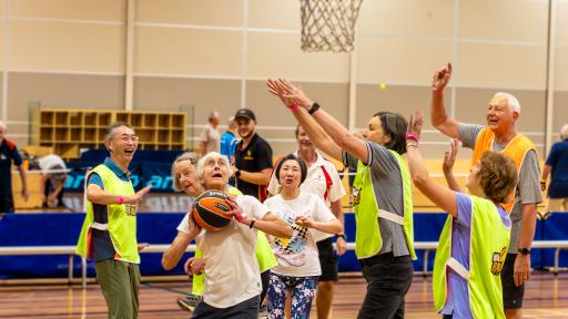 A group of older adults, half in vests, half not playing walking basketball. one person not wearing a vest has the ball and is about to try and shoot a goal.