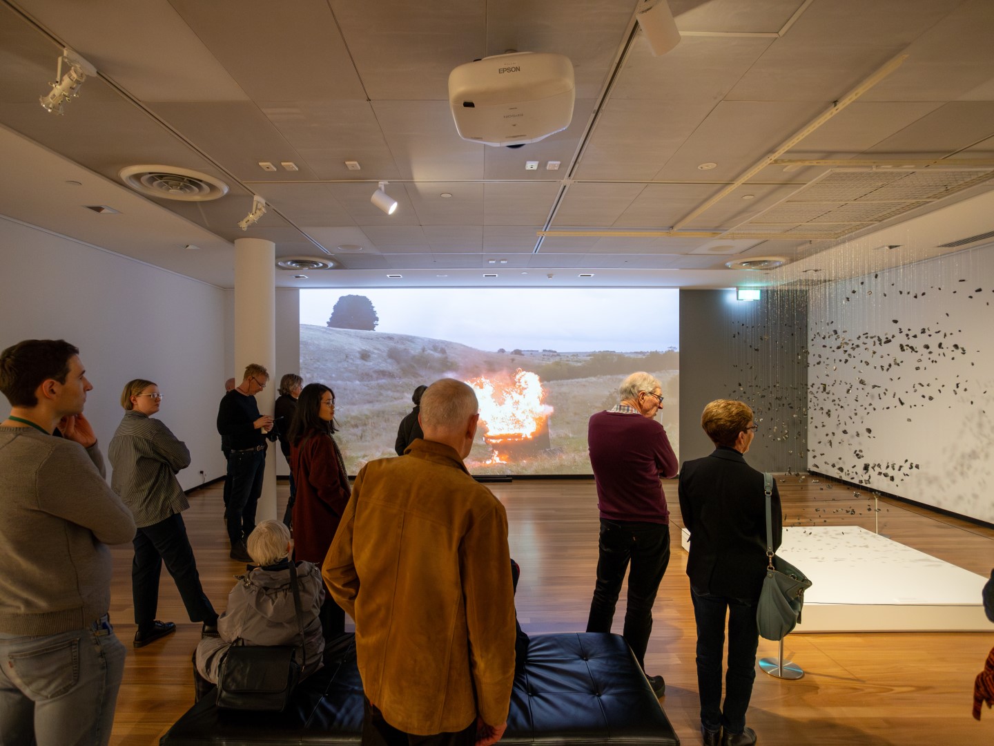 A group of people in a dimly lit gallery space looking at a projected video of fire in an empty paddock