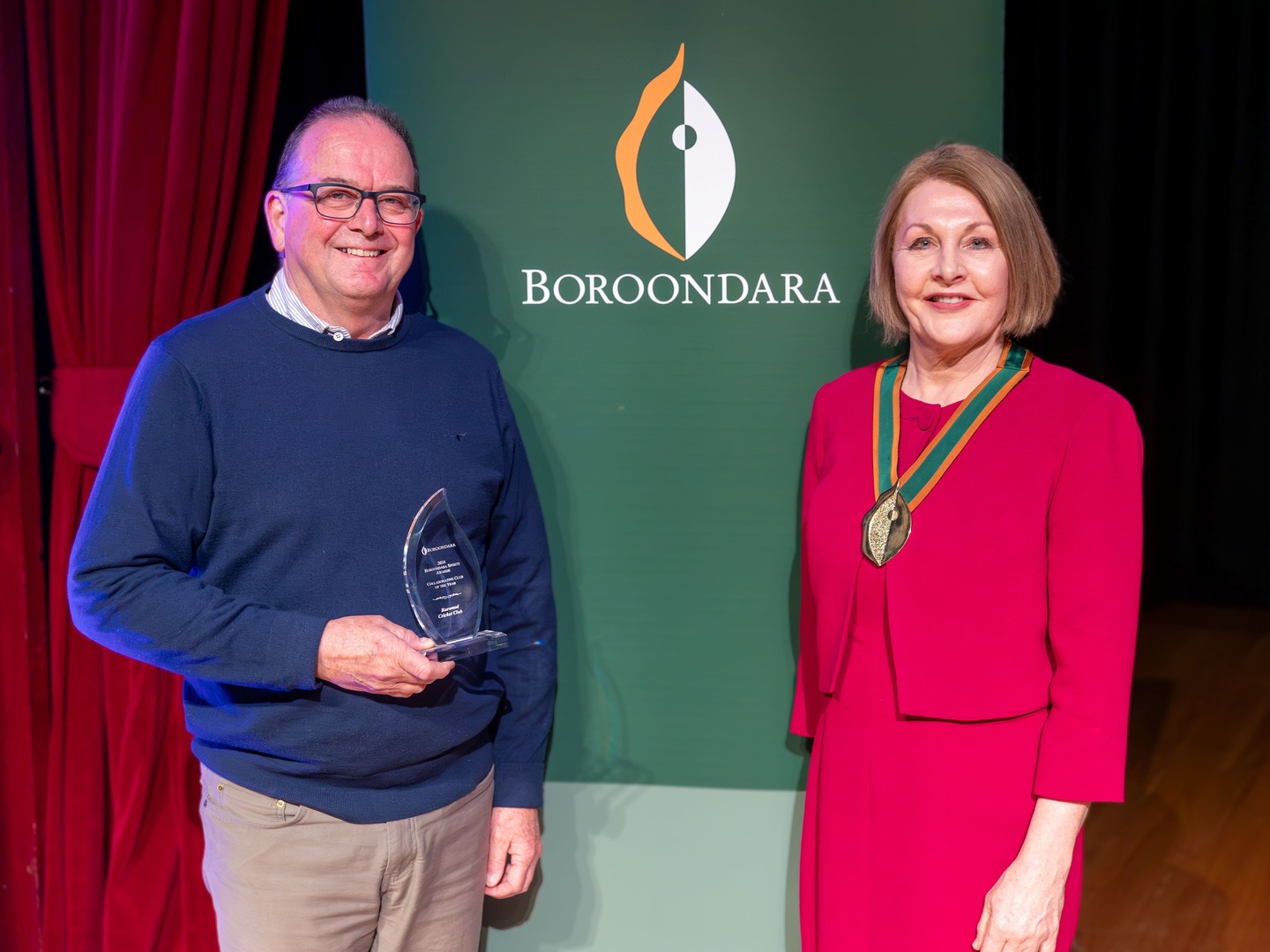 A man in a blue sweater is pictured holding a glass award. Next to him is a woman in a pink dress, adorned with a green and gold chain and a medal around her neck.  In the background, a green sign says "BOROONDARA."