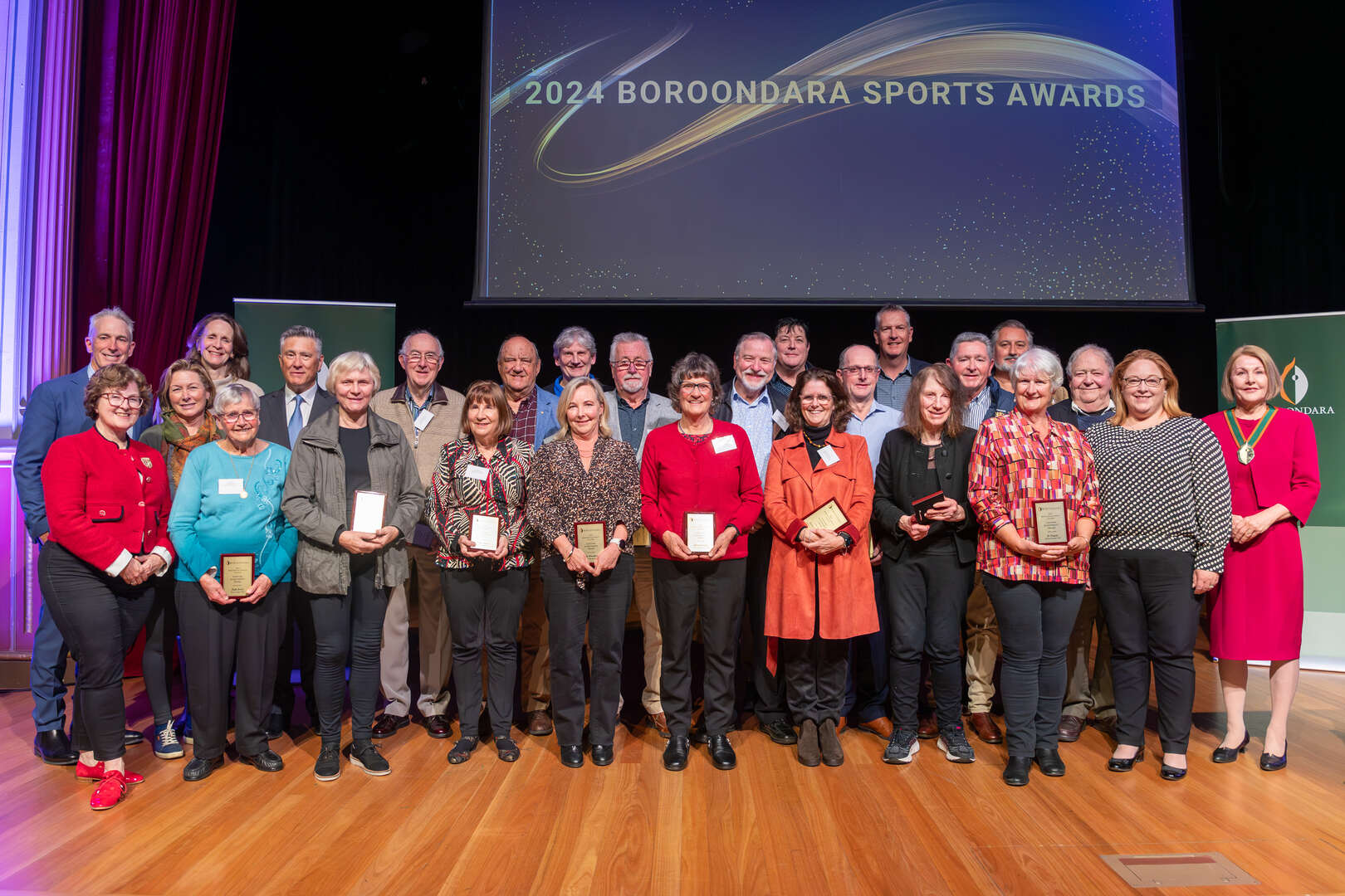 A group of 26 people, mostly older adults, are standing in a line. They are all smiling and holding small plaques. The background has a screen with the words "2024 Boroondara Sports Awards." 