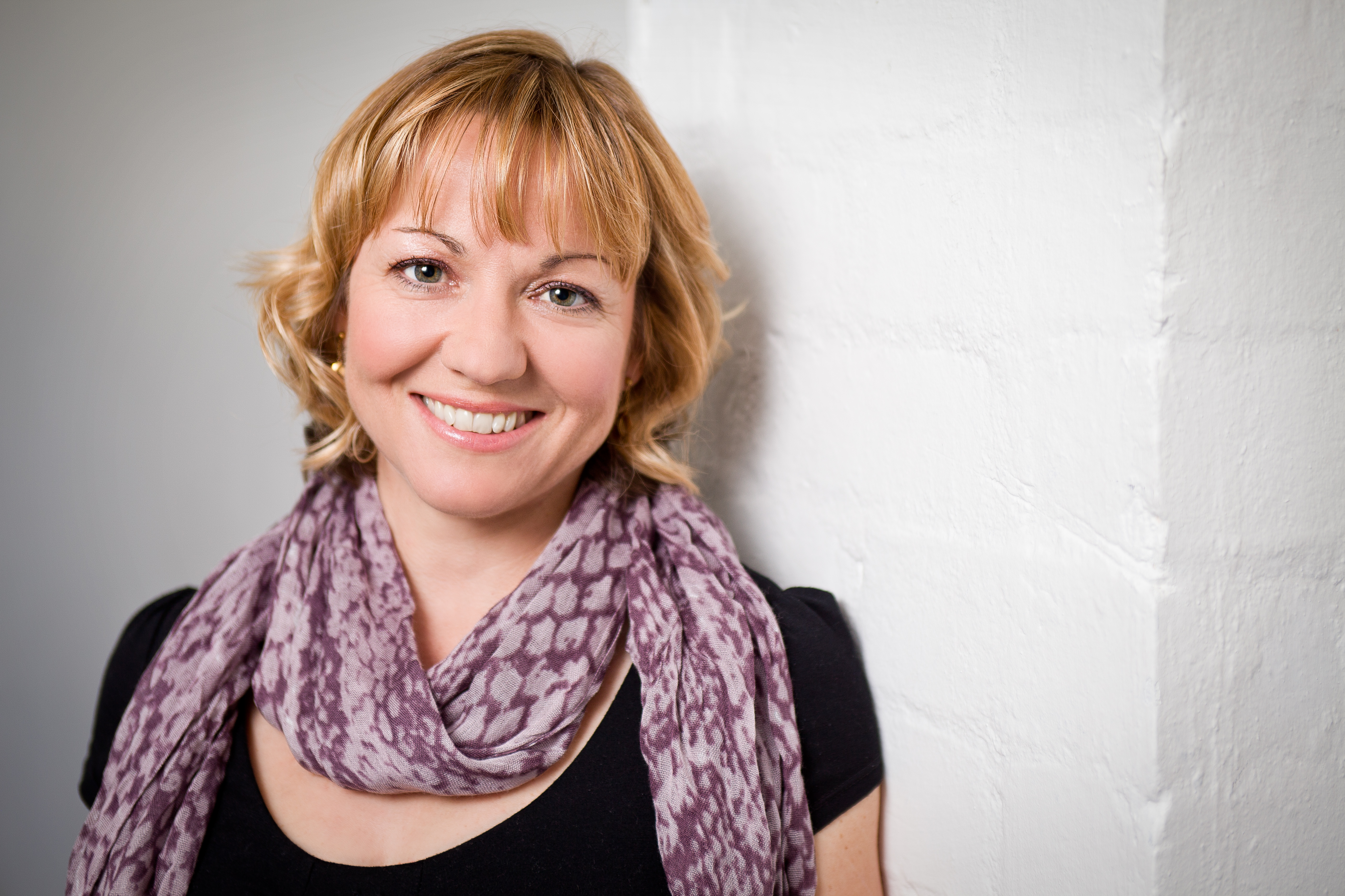 woman with short blonde hair and fringe wearing a black tshirt and purple scarf smiles