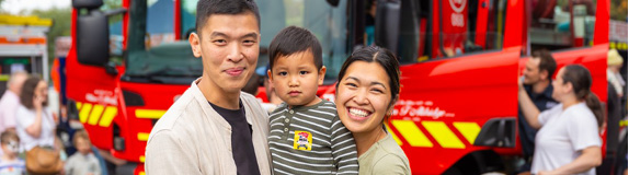 A young family of 3 standing together in front of a fire truck