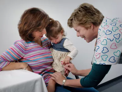 A toddler receives a jab from a nurse while sitting on a woman's lap