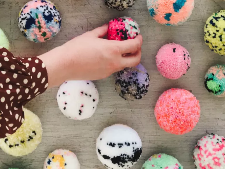 An arm reaches from the left and places a pompom on the floor, where it is surrounded by lots more pompoms.