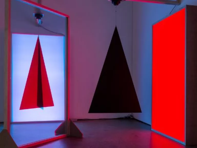 A mirror reflecting the image of a red triagle with a black line through the centre, next to a bright red lit rectange and a hanging 3D black pyramid in front of it.