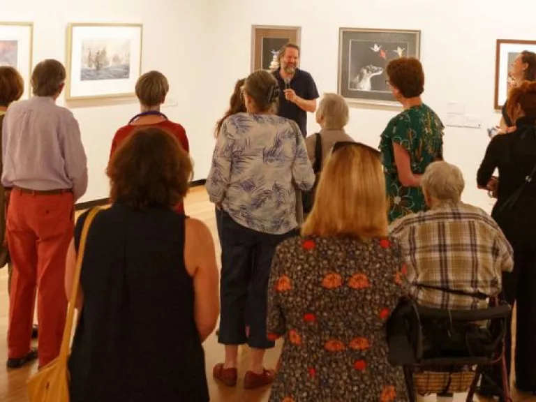 A group of people standing in an art gallery space with art handing on the walls, while listening to a talk