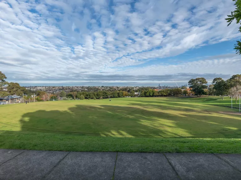 Oval-shaped grass field surrounded by two large grass and brick steps. There are trees and houses are in the distance and a footpath in the foreground.