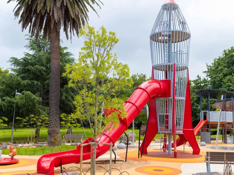 Playground with rocket-shaped climbing feature and slide, and small carousel and park bench. There are several trees in the playing area and the background.