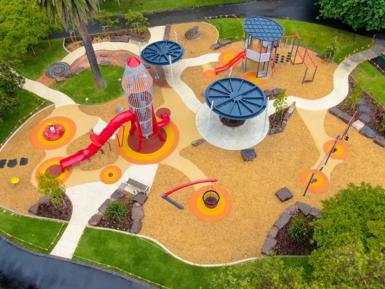 Aerial view of playground with rocket-shaped climbing feature and slide, and three other features with round tops. Grass and several large trees surround the area.