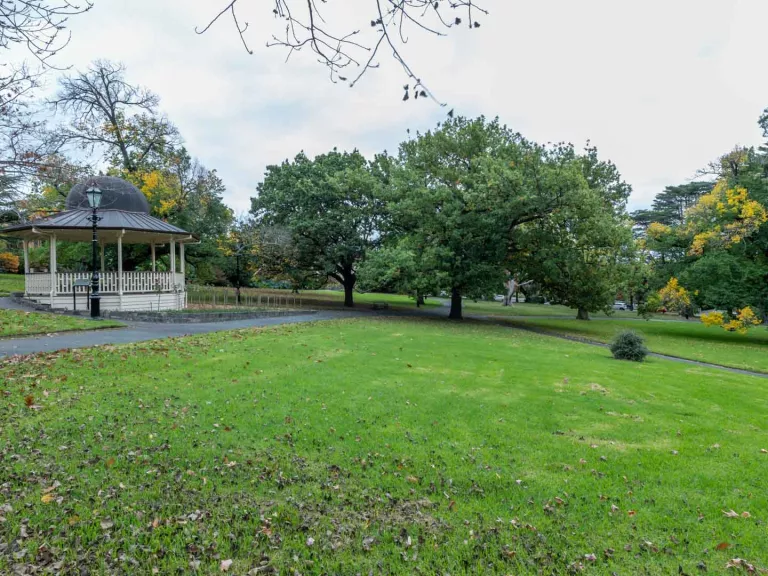 Large grass area with a left-to-right slope and a rotunda and walking path to the left. There are medium-sized trees in the background and fallen leaves in the foreground.